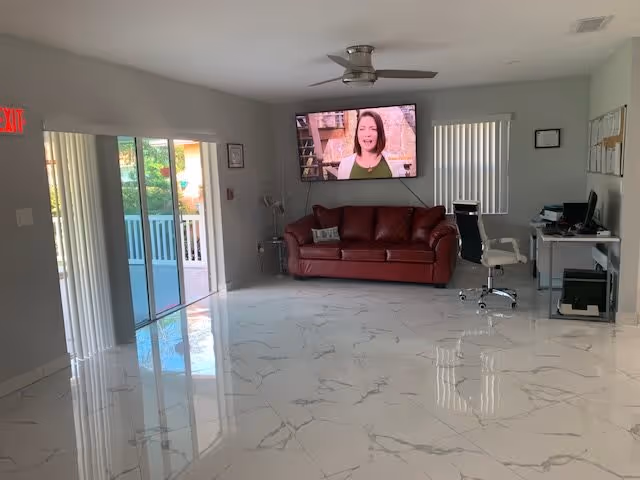 Bright common room with glossy marble tile floor, a red leather couch beneath a wall-mounted TV, a desk with chairs, and sliding glass doors to a patio.