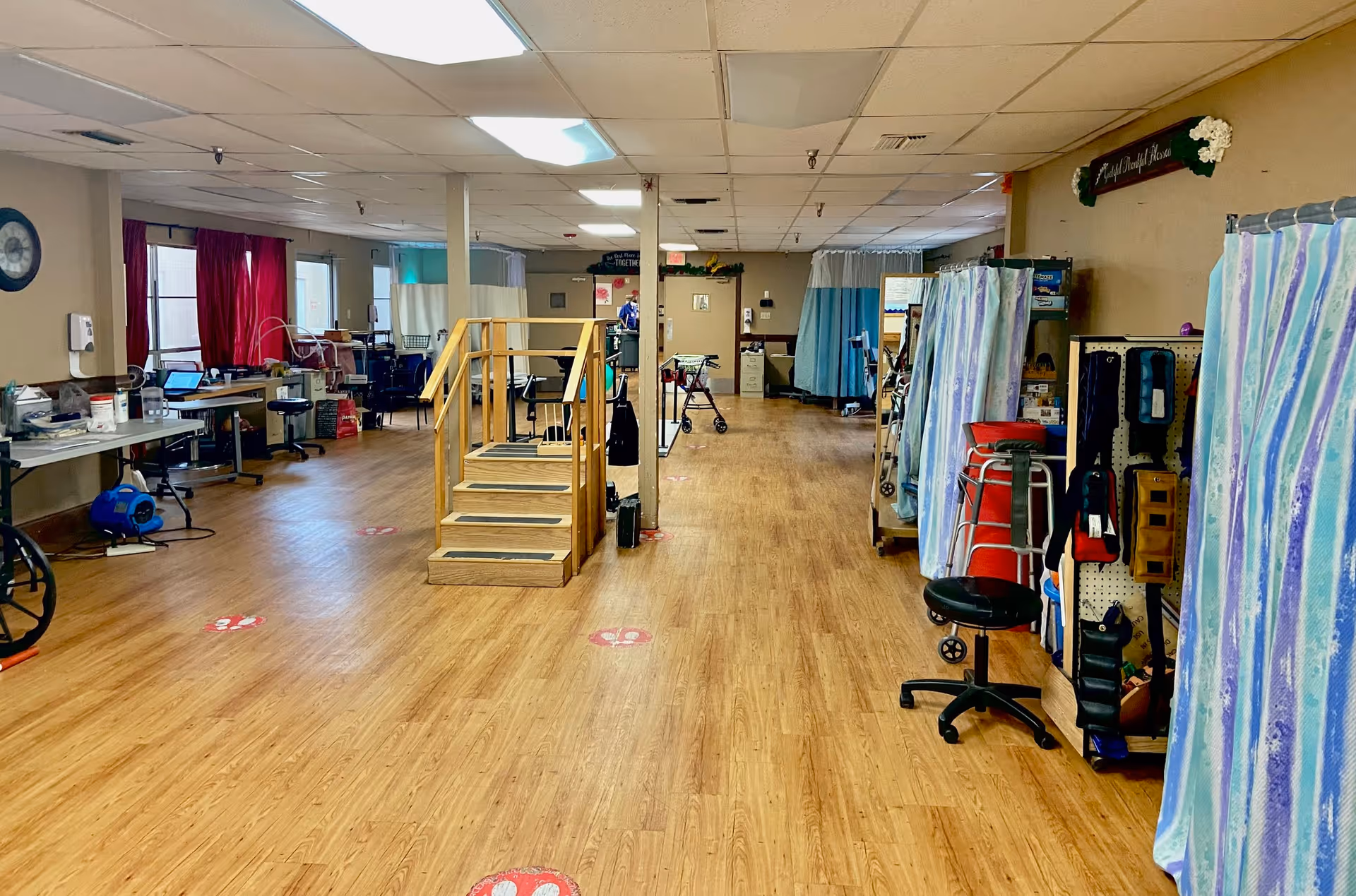 Open rehabilitation therapy room with practice stairs, mobility equipment, curtained treatment areas, and wood-look flooring.