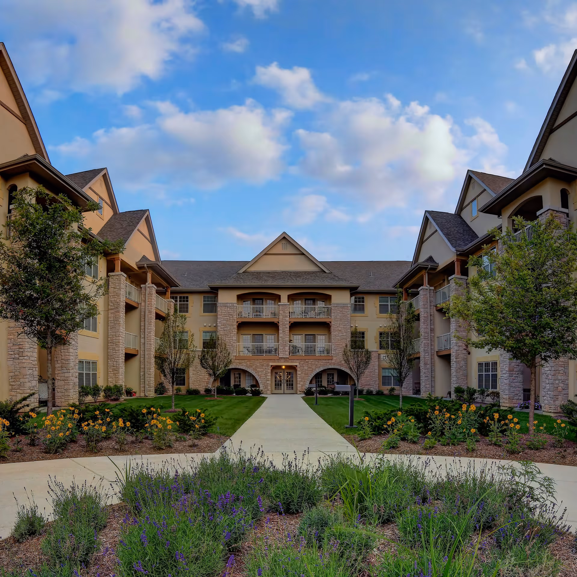 Exterior view of The Plaza at The Barrington of Carmel, showing a three-story building with stone and beige siding, balconies, and a landscaped garden with flowers and trees under a partly cloudy blue sky.
