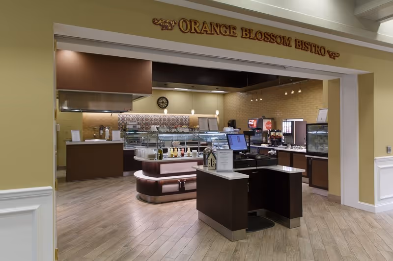Interior view of Orange Blossom Bistro featuring a serving counter with various food and drink dispensers, a cash register, and a tiled backsplash behind the counter. The area is well-lit with pendant lights and has a wood-patterned floor.