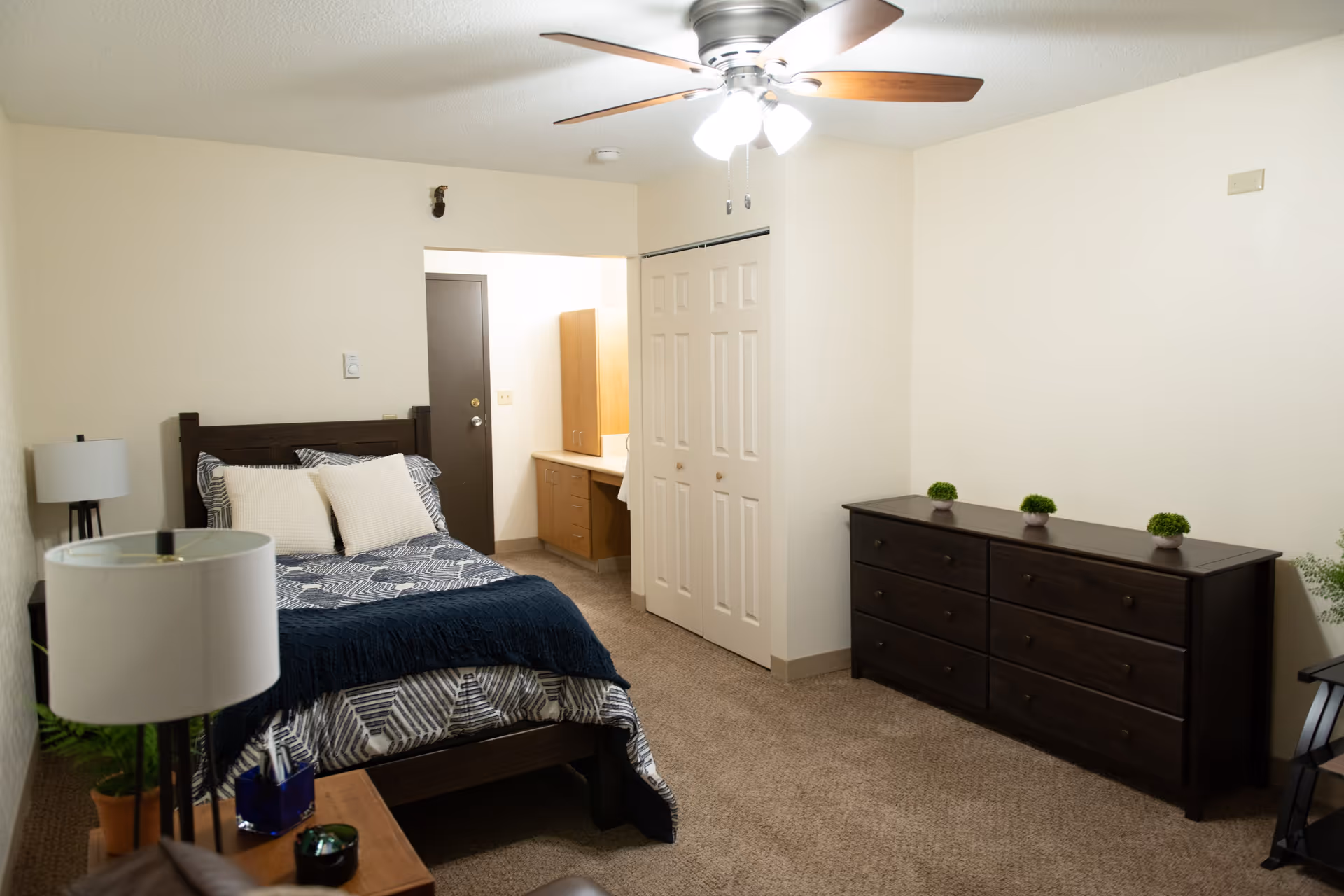 A bedroom with a bed featuring patterned bedding and two pillows, a ceiling fan with lights, a dark wooden dresser with three small potted plants on top, a nightstand with a lamp and some small items, a closet with white double doors, and a small vanity area with cabinets and a mirror in the background.