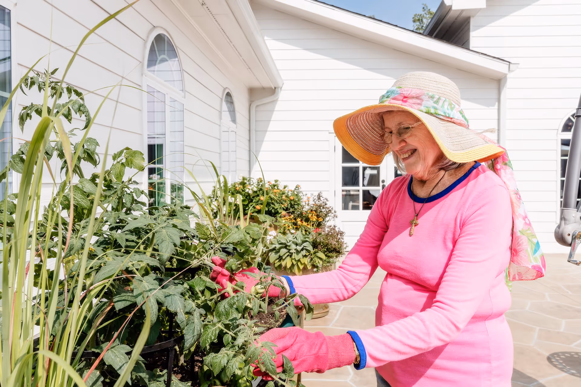 An elderly woman wearing a large sun hat, pink long-sleeve shirt, and pink gardening gloves is tending to plants in a garden area outside a white building with arched windows.