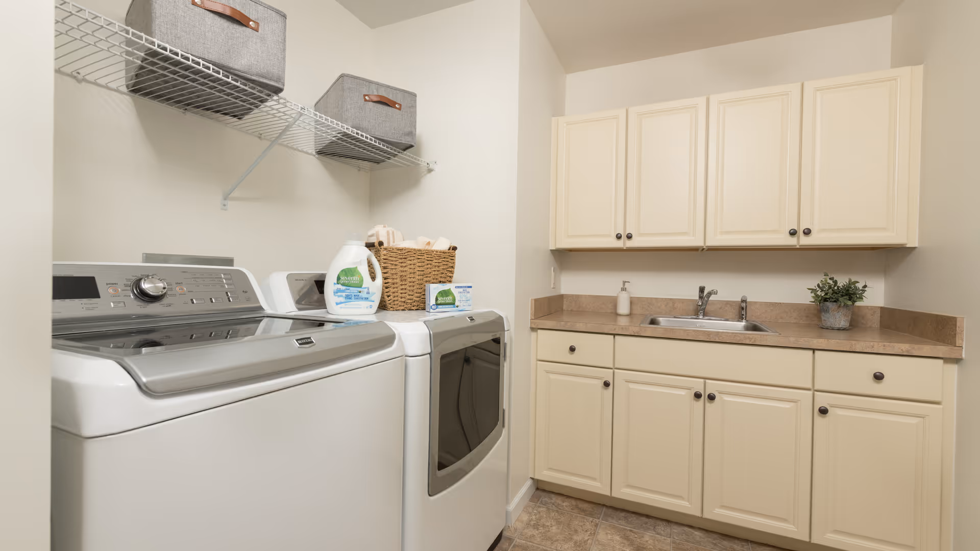 Laundry room with side-by-side washer and dryer, a countertop sink, cream cabinets, and wire shelving holding storage baskets.
