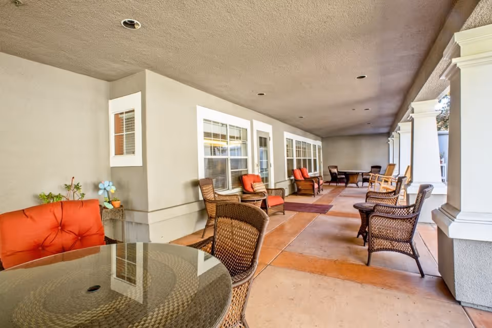 Covered outdoor patio area with multiple wicker chairs and tables, some with red cushions, adjacent to a building with white-framed windows and columns.