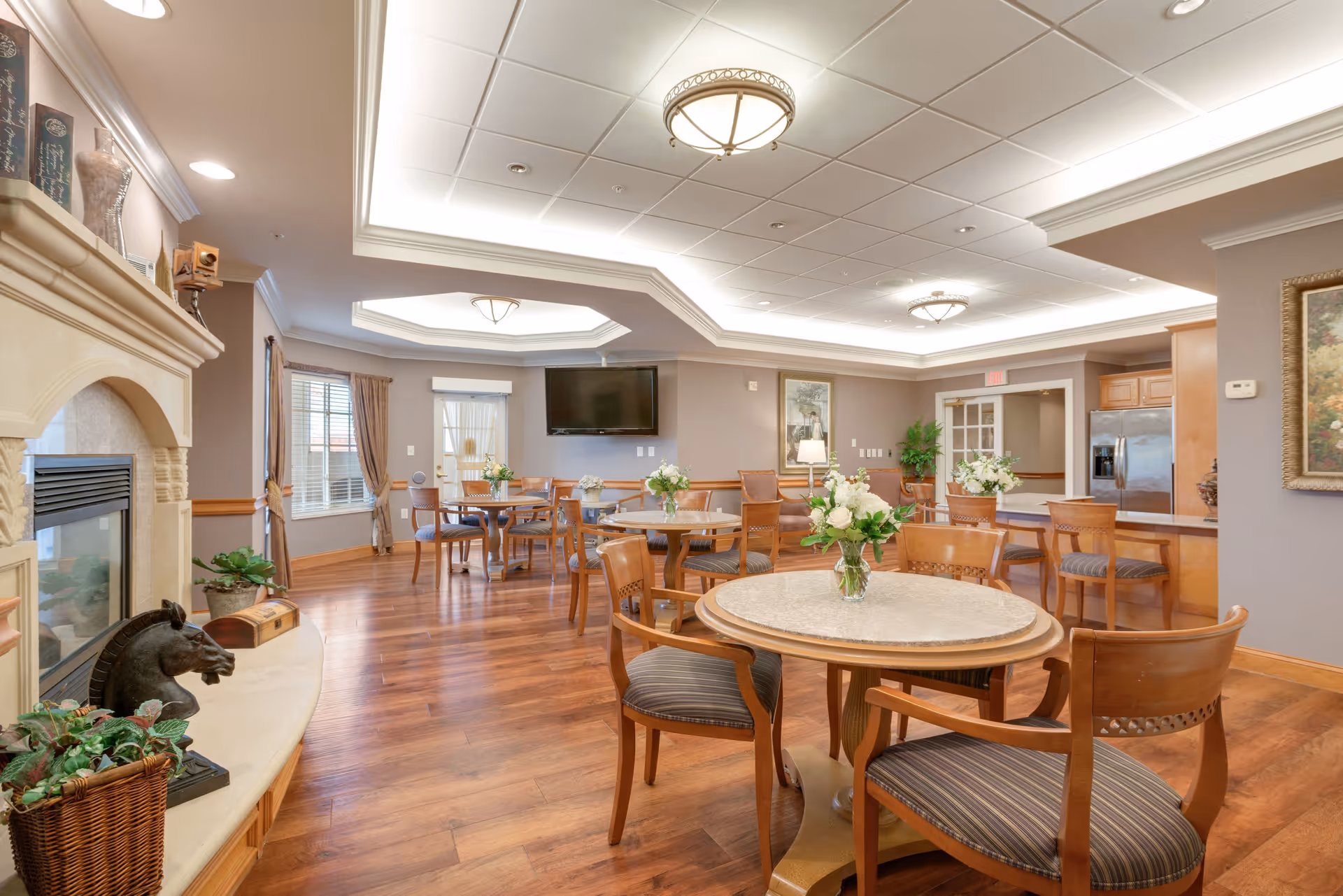 A bright and spacious senior living common area with round tables and wooden chairs arranged neatly. The room features a fireplace on the left, a flat-screen TV mounted on the far wall, and a kitchen area with a stainless steel refrigerator on the right. The ceiling has recessed lighting and decorative light fixtures, and there are floral centerpieces on the tables. Large windows with curtains allow natural light to enter the room.