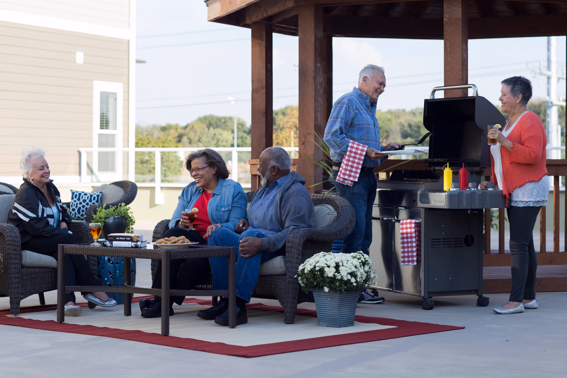 Five elderly people enjoying an outdoor gathering on a patio. Three are seated on wicker chairs around a coffee table with drinks and snacks, while two others stand near a barbecue grill, smiling and talking. The setting includes a red-bordered outdoor rug, potted flowers, and a wooden pergola overhead.