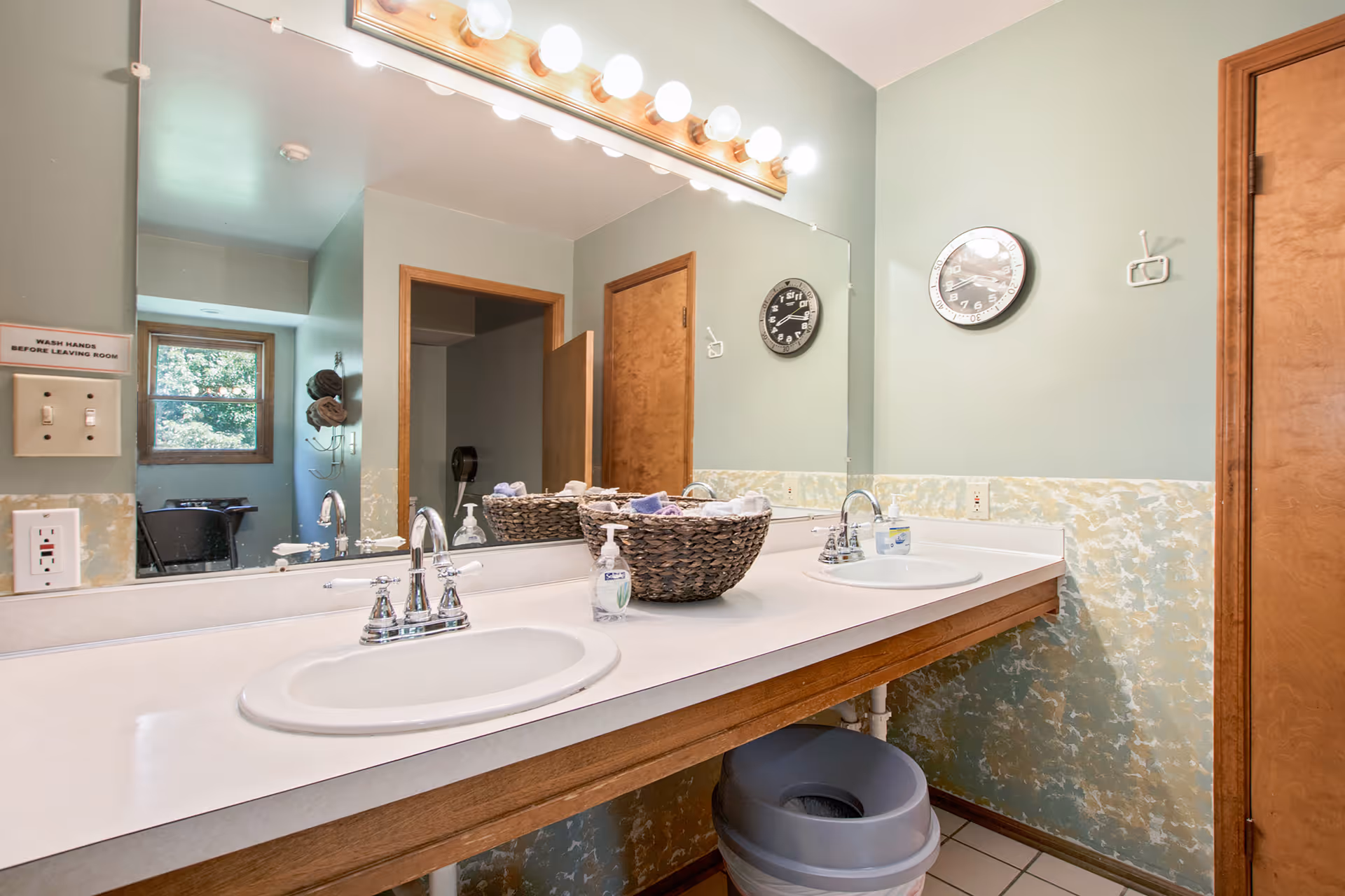 A bathroom with a double sink vanity featuring two chrome faucets, a large mirror with lights above, a basket with towels, a soap dispenser, a clock on the wall, and wooden doors. The walls are painted light green with a textured lower half, and there is a trash can under the sink.