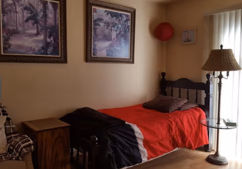 A bedroom with a bed covered by a red blanket, nightstand, lamp, and framed wall art.