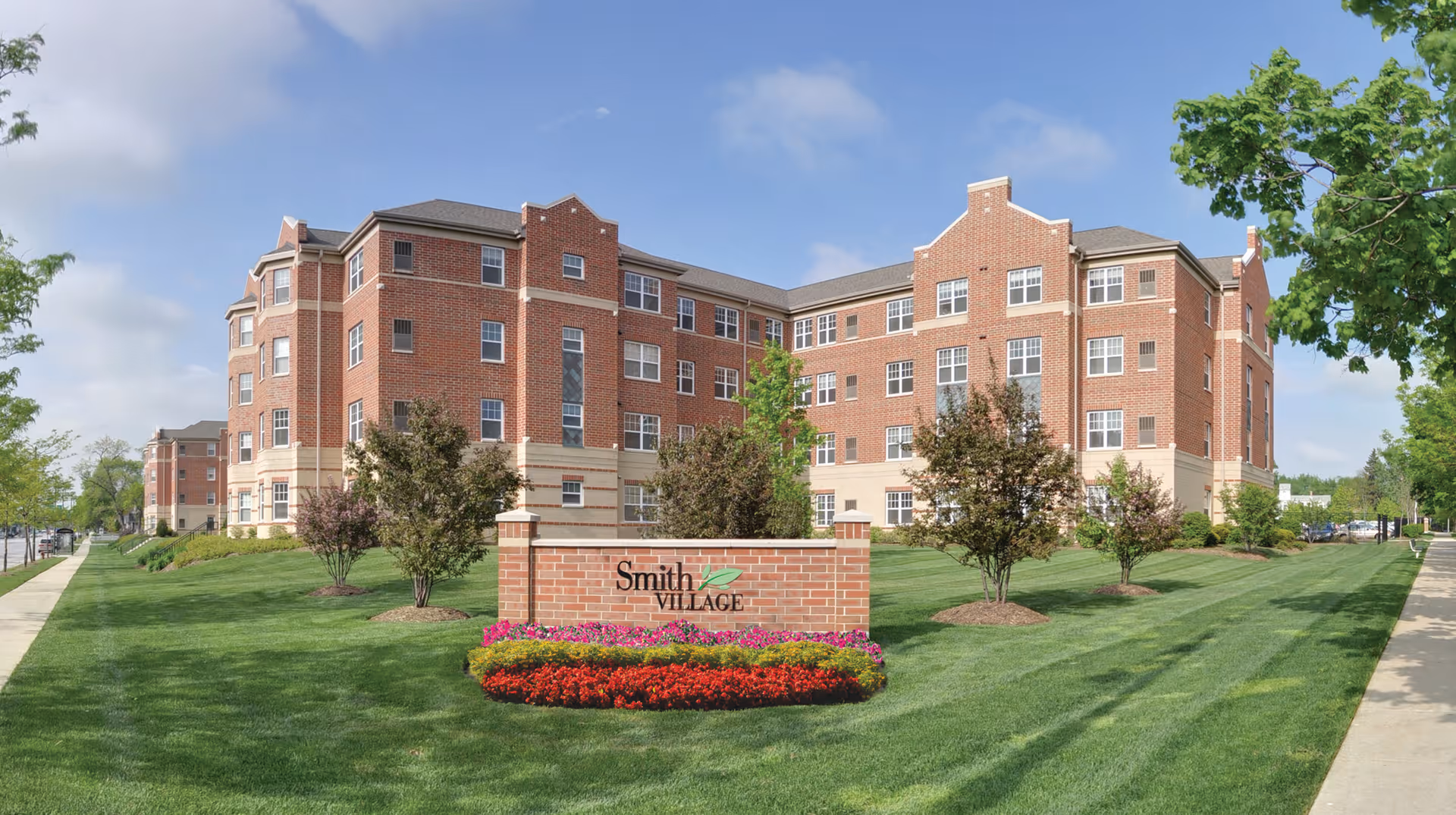 Exterior view of a large, multi-story brick building with multiple windows, surrounded by green lawns, trees, and flower beds. In front of the building is a brick sign with the text 'Smith Village'. The sky is partly cloudy.