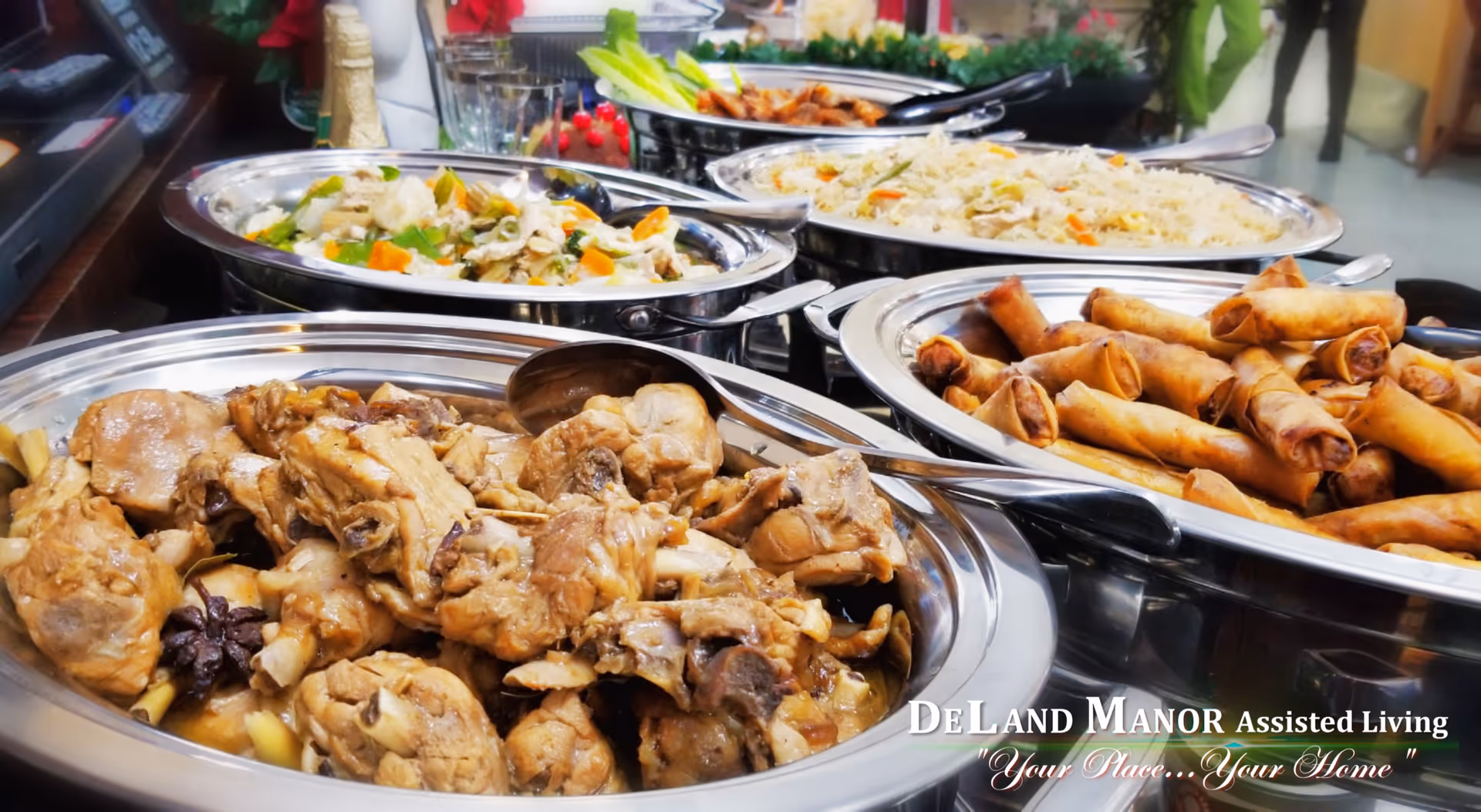 Buffet-style serving trays filled with various cooked dishes including chicken, spring rolls, rice, and mixed vegetables, arranged on a table in a dining area.