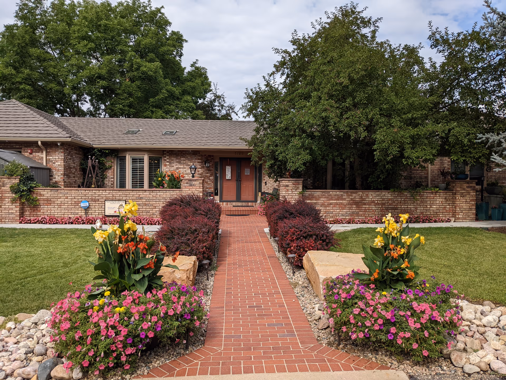Front exterior view of Monarch Greens Assisted Living facility showing a brick building with a tiled roof, a central pathway lined with red bushes and colorful flower beds on either side, green lawn, and large trees in the background under a partly cloudy sky.