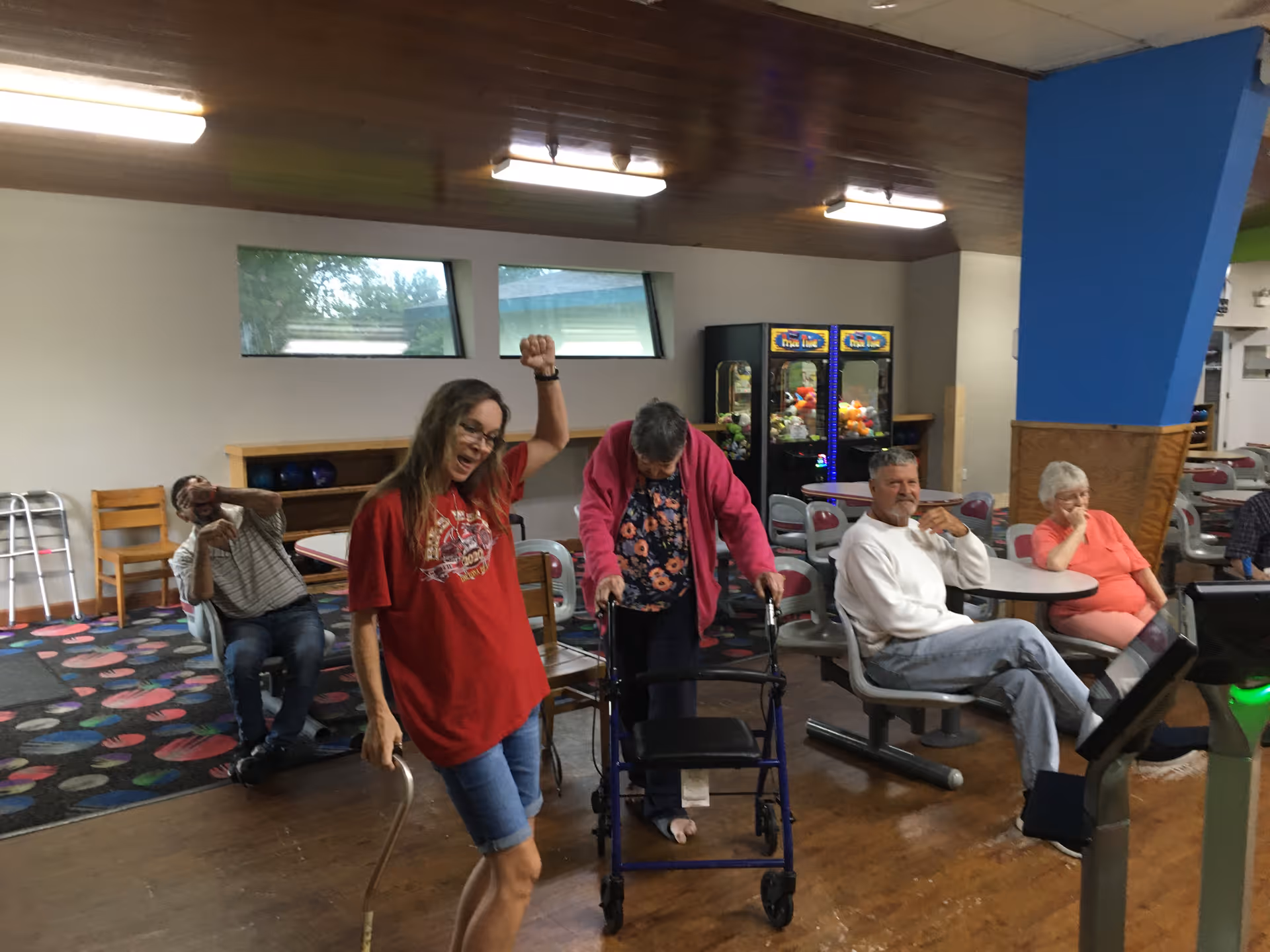 A group of elderly people and a younger woman are in a recreational room with bowling lanes. The younger woman in a red shirt and shorts is standing and smiling with her arm raised. An elderly woman with a walker is standing nearby. Three other elderly individuals are seated at tables and chairs, with two arcade claw machines visible in the background. The room has a wooden floor and ceiling with fluorescent lights.