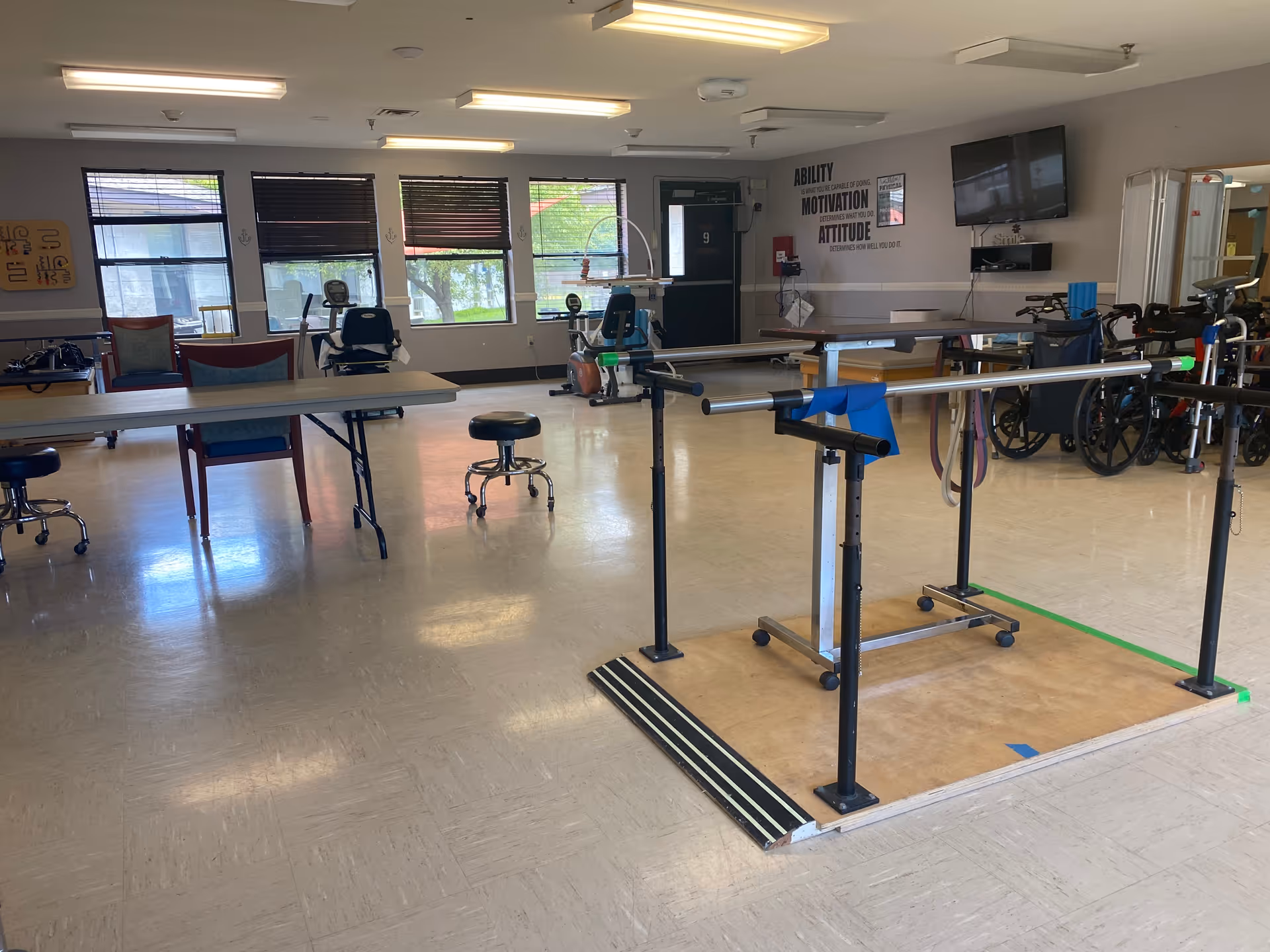 Spacious interior therapy room with parallel walking bars, wheelchairs, tables and chairs, and windows.