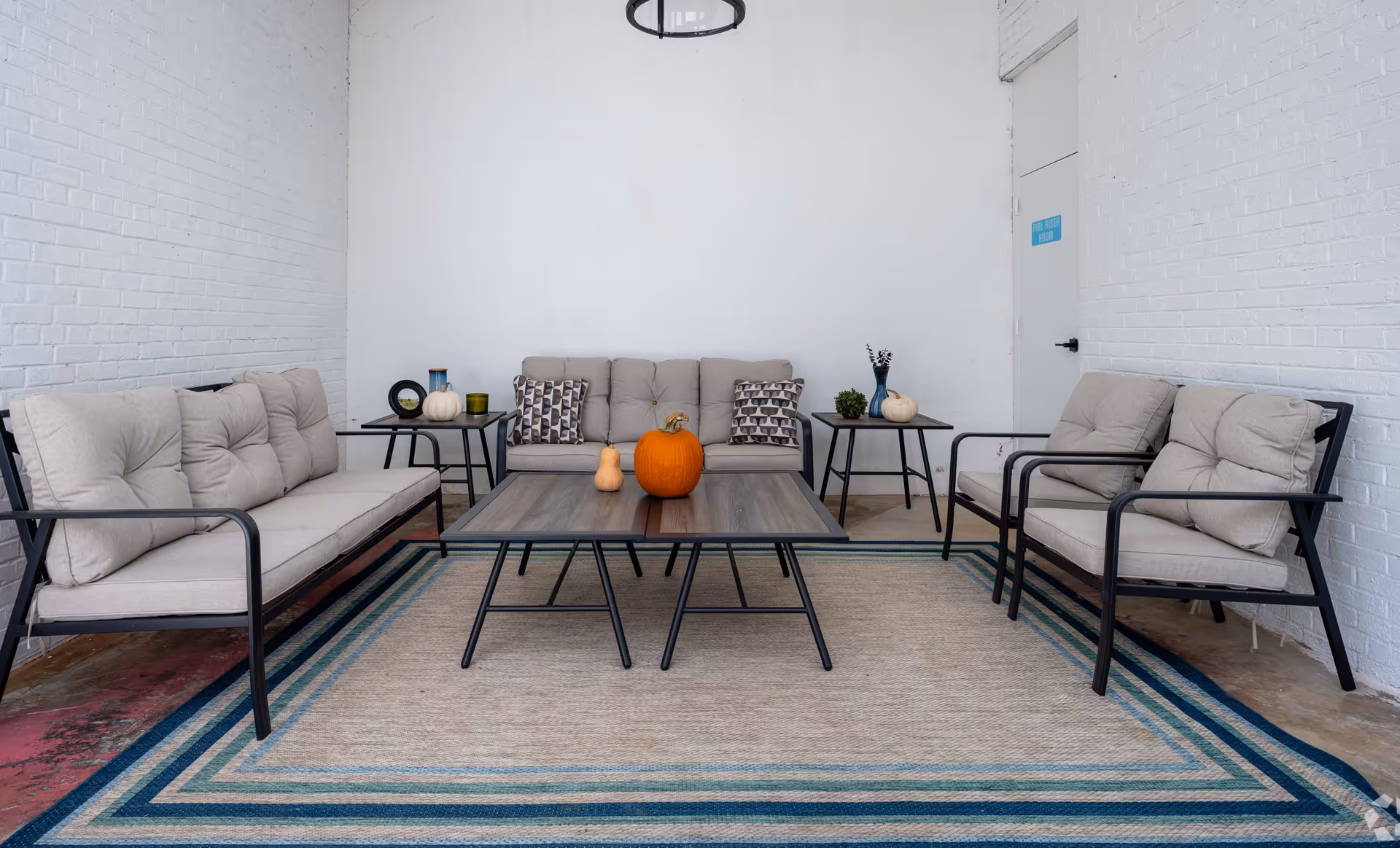 A cozy seating area with three cushioned sofas arranged around two wooden coffee tables. The tables have pumpkins and decorative items on them. The room has white brick walls and a large rug with blue and beige stripes on the floor.