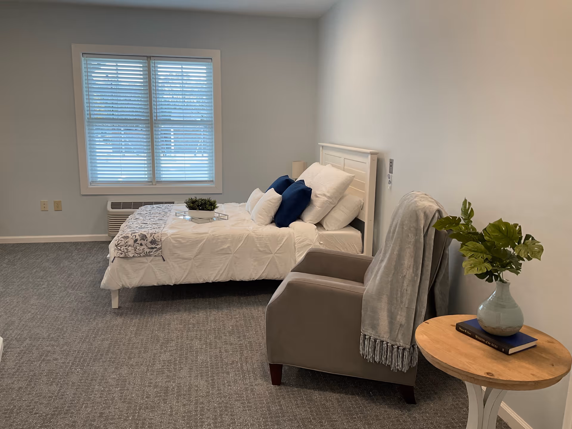 A neatly made bed with white and blue pillows and a white headboard is positioned against a light-colored wall. Next to the bed is a gray armchair draped with a gray blanket. A small round wooden table beside the chair holds a blue vase with green leaves and a book. A window with white blinds is on the wall behind the bed, and the floor is carpeted in gray.