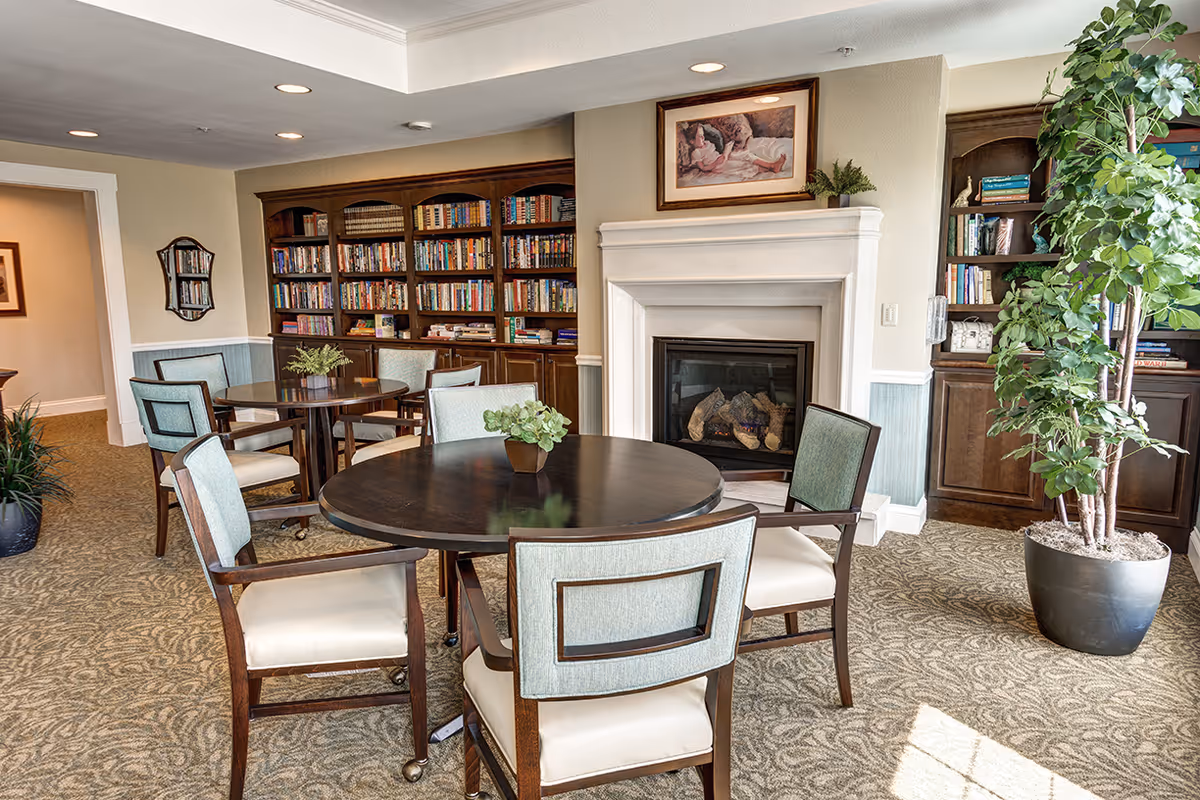 A cozy senior living common area with round wooden tables and cushioned chairs. There is a fireplace with a framed painting above it, built-in wooden bookshelves filled with books, and a large potted plant near the window letting in natural light.