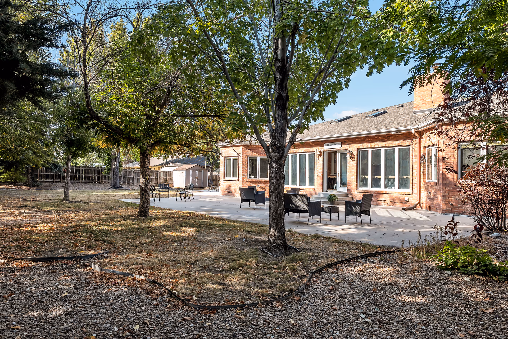 Outdoor patio area at a senior living facility with several chairs and tables arranged on a concrete surface. The patio is adjacent to a brick building with large windows and a door. Trees and a fenced yard surround the patio, with a shed visible in the background under a clear blue sky.