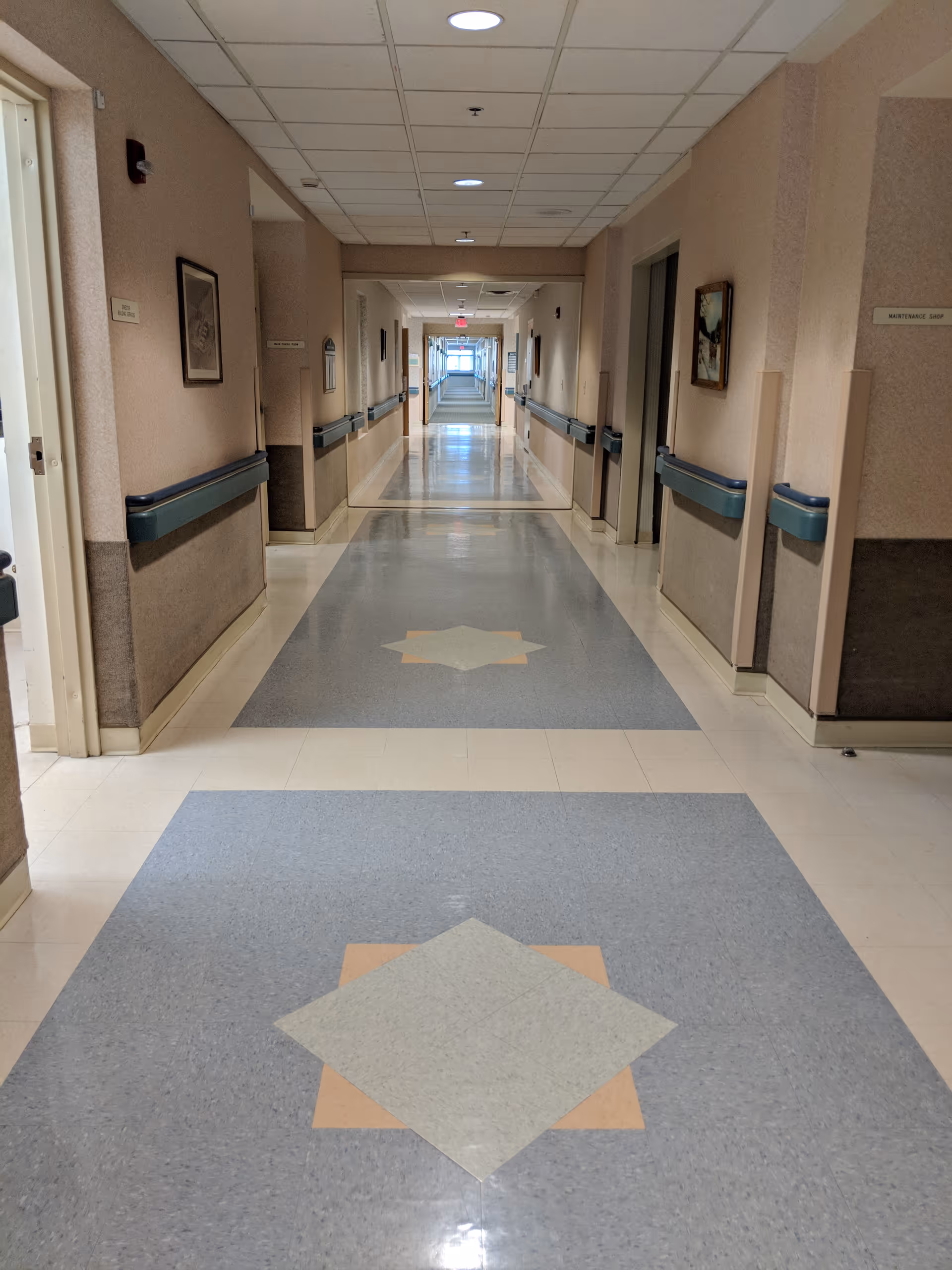 A long, clean hallway in a nursing and rehabilitation center with patterned tile flooring, beige walls, handrails on both sides, framed pictures on the walls, and ceiling lights. Doors and signs are visible along the corridor.