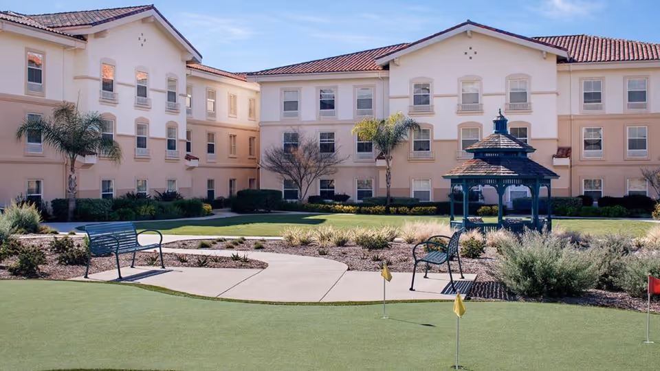 Outdoor courtyard area of Belmont Village Senior Living Sabre Springs featuring a putting green with small flags, a paved walkway, two metal benches, landscaped bushes and plants, palm trees, and a gazebo in front of a three-story beige building with multiple windows under a clear blue sky.