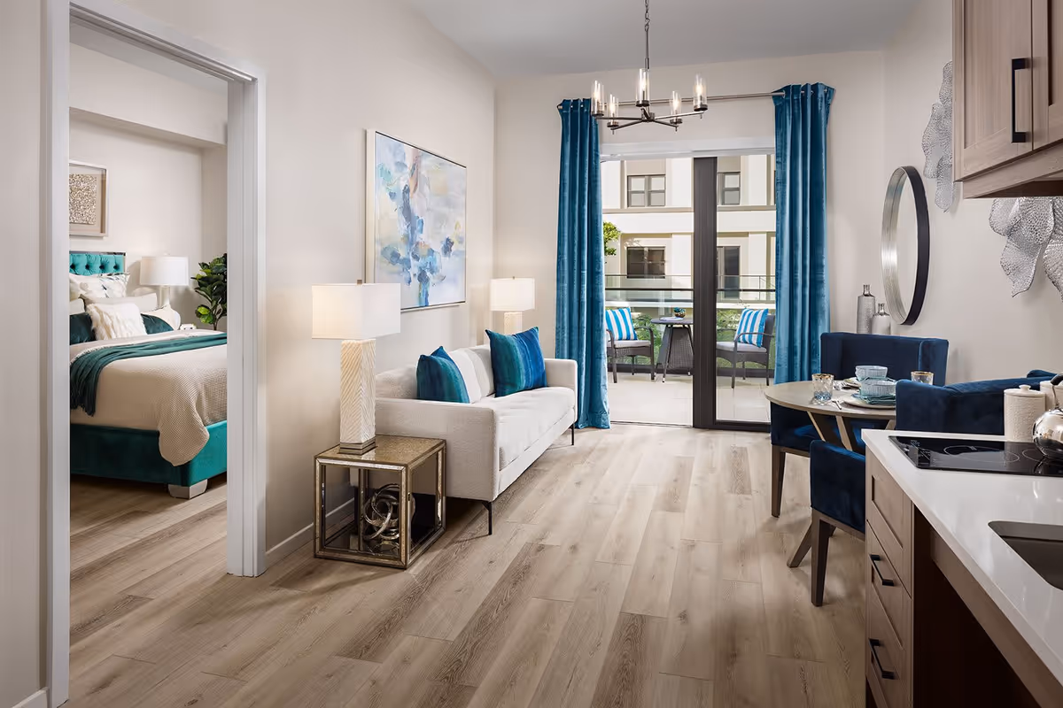 A modern senior living apartment interior featuring a small living room with a white sofa and blue cushions, a side table with a lamp, and a large abstract painting on the wall. To the right is a dining area with a round table and blue upholstered chairs. The kitchen area with wooden cabinets and a white countertop is partially visible. Large glass sliding doors with blue curtains open to a balcony with two chairs and a small table. A bedroom with a teal upholstered bed and bedside lamp is visible through an open doorway on the left.