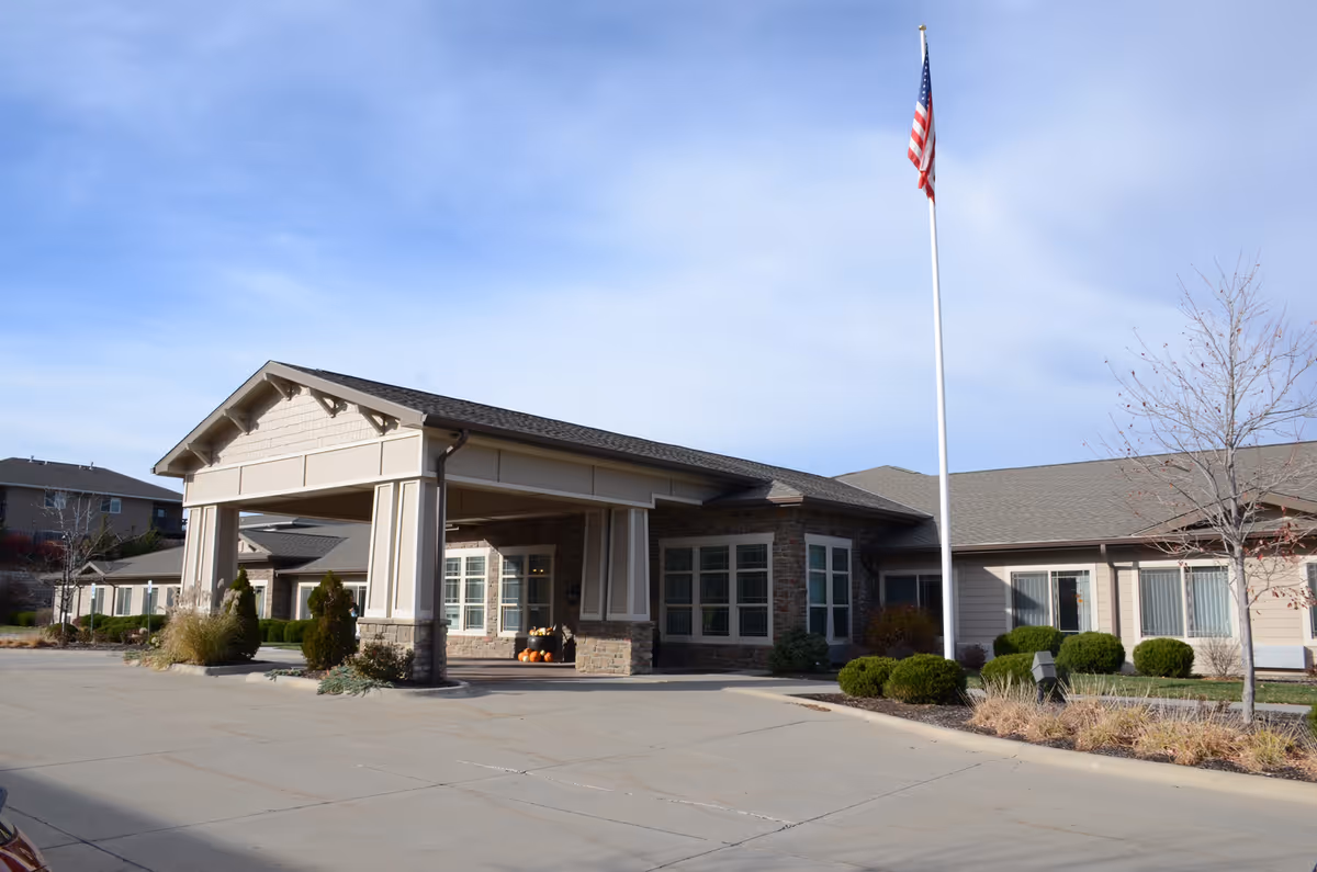 Front entrance of a single-story senior living building with a covered porte-cochere, landscaping, and a tall American flag.