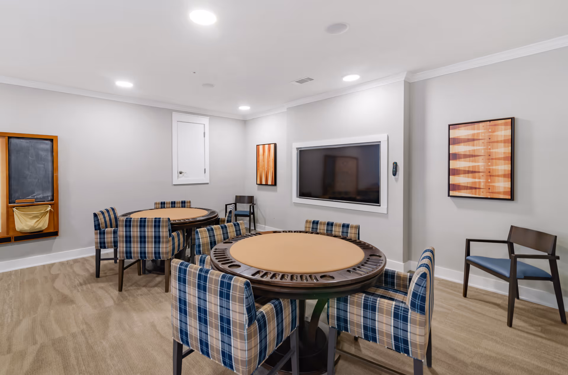 Game room with round card tables surrounded by plaid chairs, a wall-mounted TV, and framed artwork.