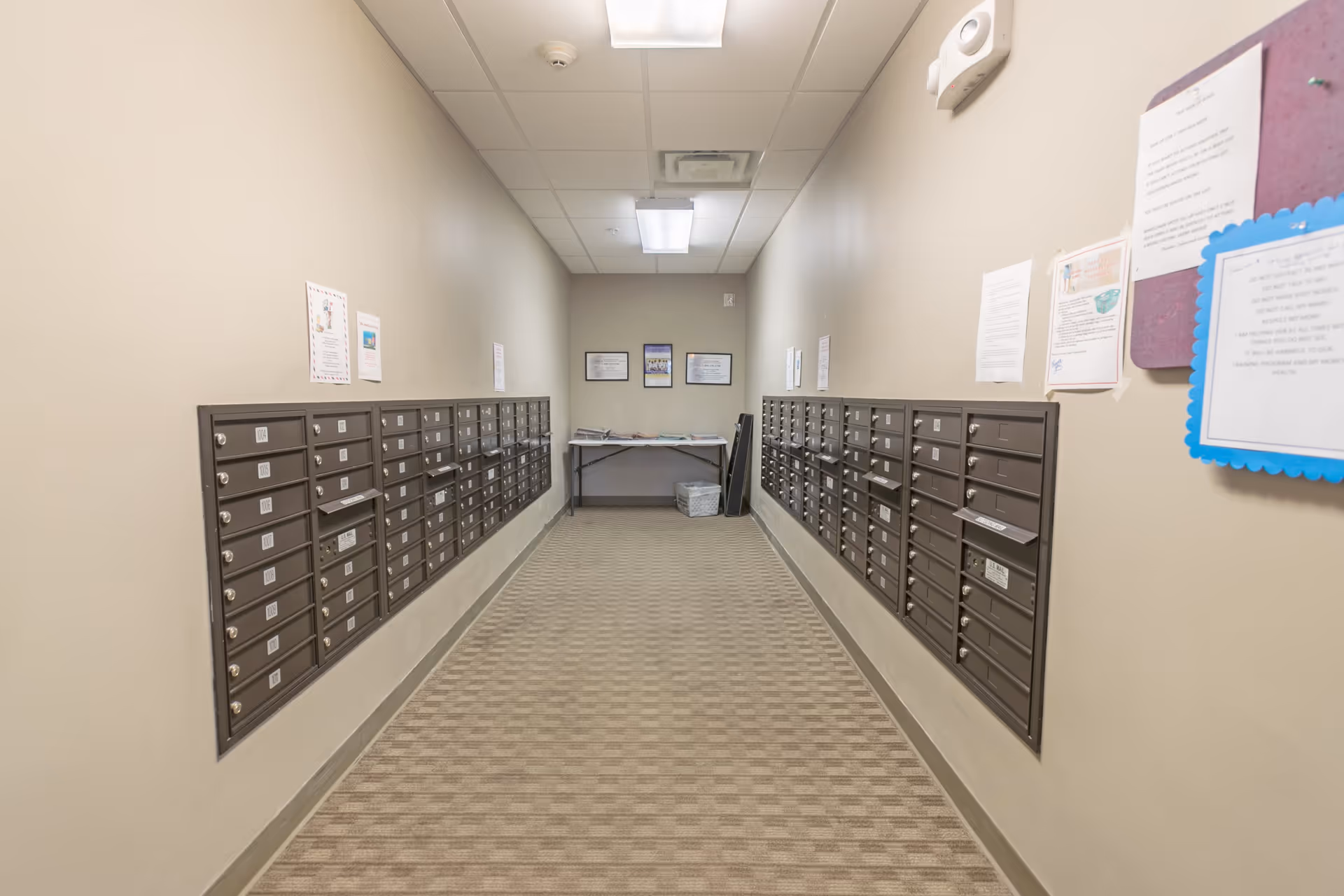 A narrow hallway with rows of mailboxes mounted on both walls. The floor is carpeted with a checkered pattern, and the walls are beige with various notices and papers pinned on the right side. At the end of the hallway, there is a table with newspapers and a wastebasket underneath. The ceiling has fluorescent lighting panels.