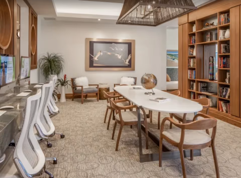 Bright communal interior with a long white table surrounded by wooden chairs, computers and office chairs along the left, and bookshelves on the right.