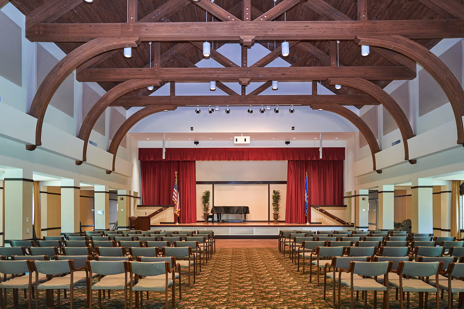 Interior view of an auditorium or event hall with rows of chairs facing a stage with a grand piano, two potted plants, and red curtains. The ceiling features large wooden beams and multiple ceiling lights.