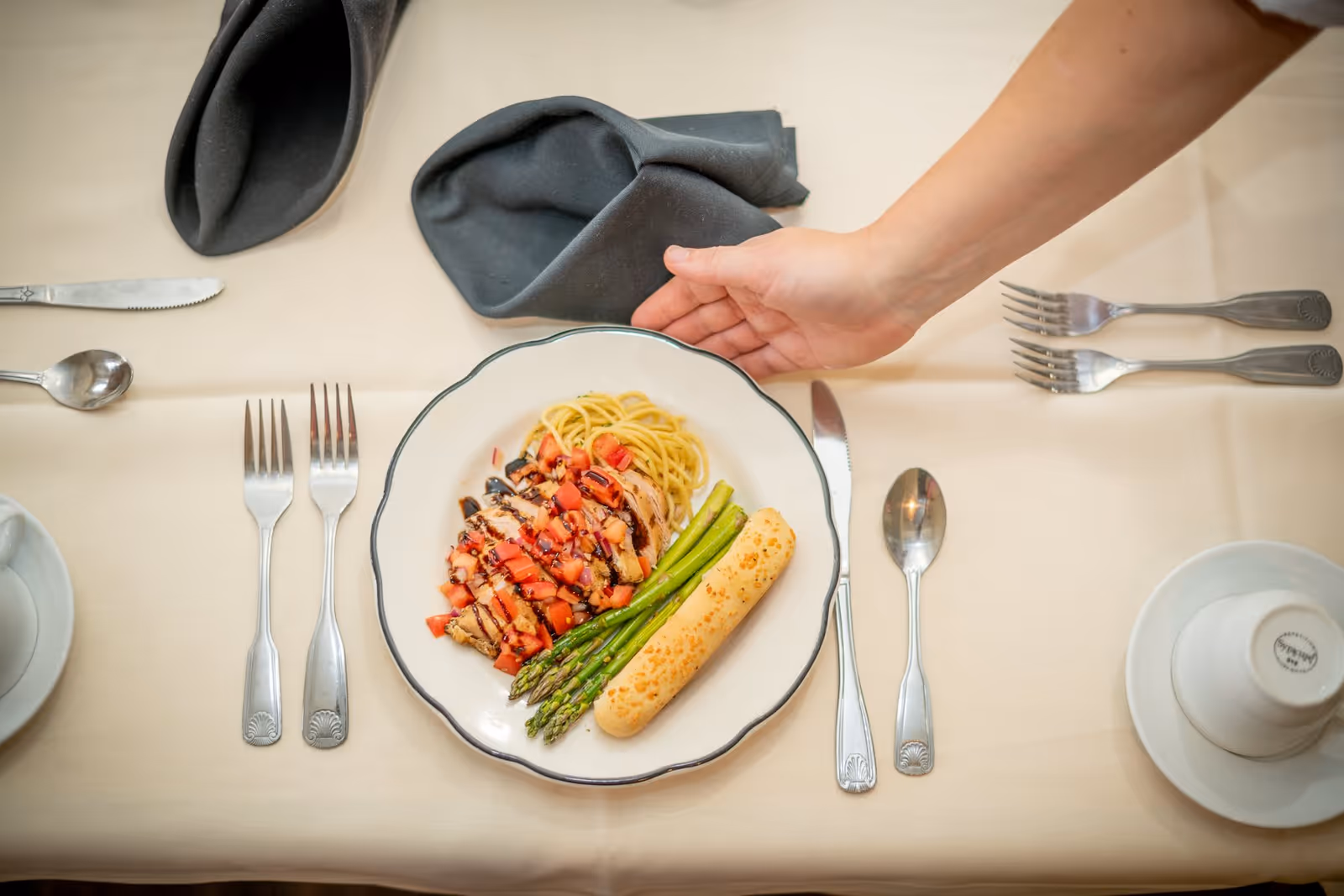 A plate of food on a table with spaghetti, grilled chicken topped with diced tomatoes and balsamic glaze, steamed asparagus, and a breadstick. A hand is reaching towards the plate. The table is set with silverware, a cup and saucer, and a folded dark napkin on a light-colored tablecloth.