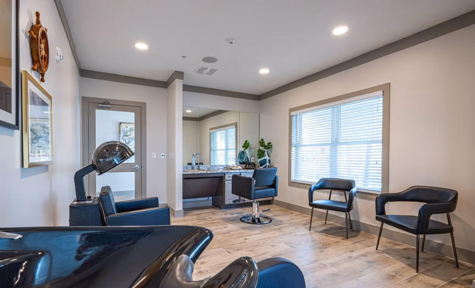 Interior view of a modern hair salon area in a senior living facility with a hair washing station, salon chairs, a large mirror, a countertop with a sink, and two black chairs near a window with blinds.