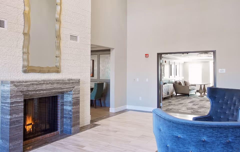 Bright communal lounge with a marble fireplace, decorative mirror, blue upholstered chairs, and a view into an adjoining seating room.
