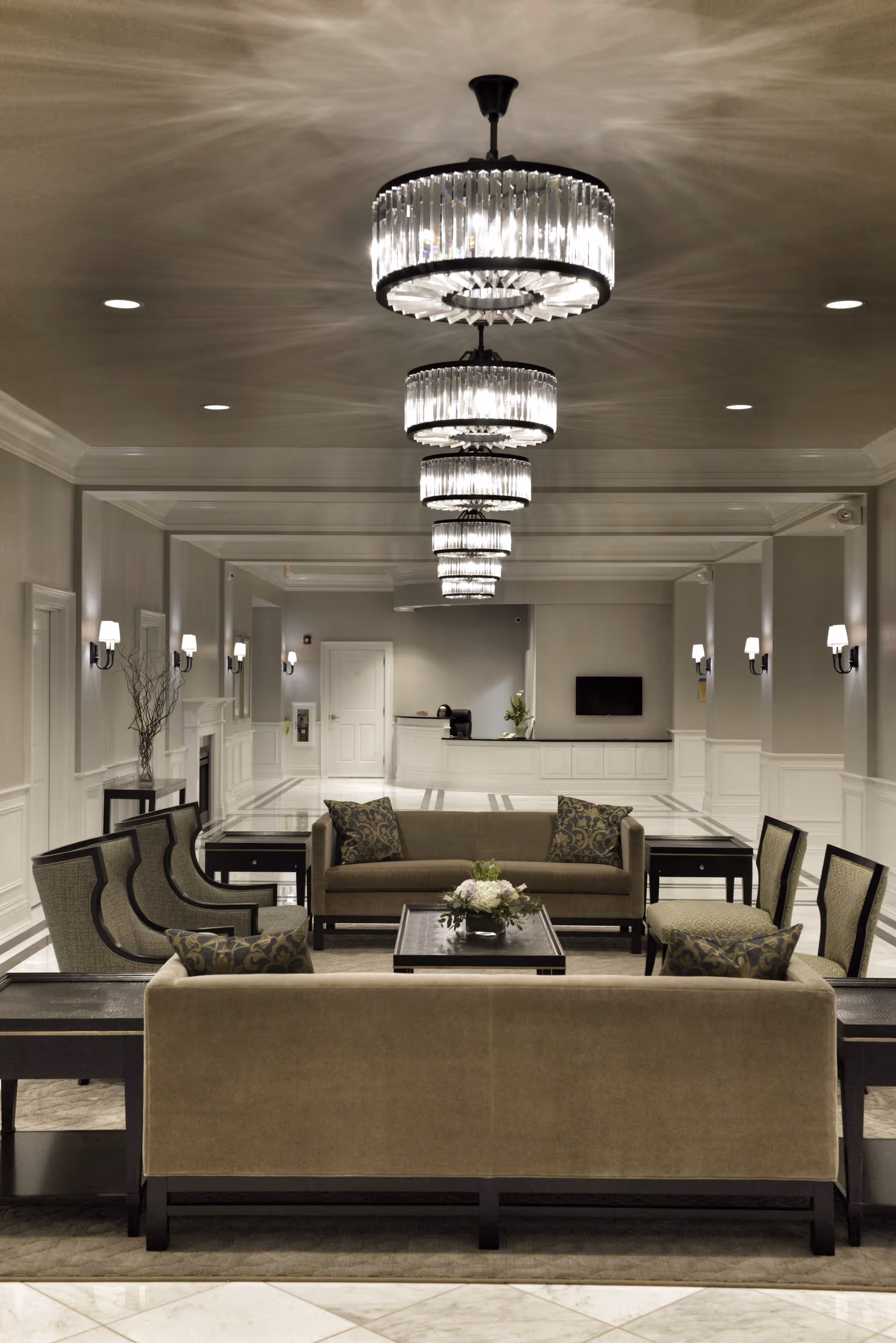 A spacious and elegant lounge area with beige sofas and armchairs arranged around a dark wooden coffee table with a floral centerpiece. The room features multiple crystal chandeliers hanging from the ceiling, wall sconces, and a reception desk with a TV mounted on the wall behind it. The decor is neutral with light-colored walls and marble flooring.
