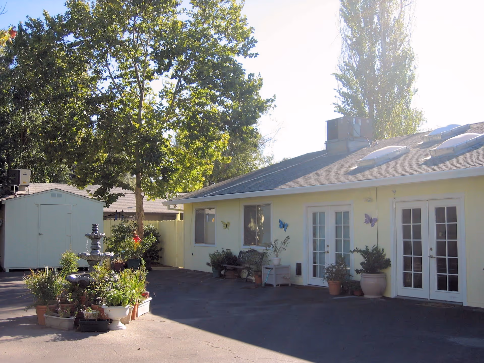 Single-story yellow building exterior with French doors, potted plants, a fountain, and a storage shed under trees.