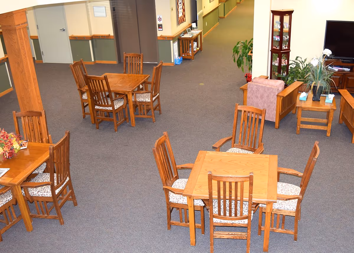 Interior view of a senior living facility common area with multiple wooden tables and chairs arranged on a carpeted floor. There are plants, a television, and a small cabinet with decorative items in the background. The walls are painted in neutral tones with wooden trim and handrails along the hallway.