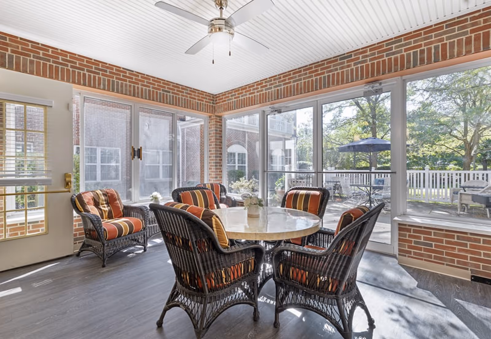 Sunlit enclosed brick sunroom with a round dining table surrounded by wicker chairs and large windows overlooking an outdoor patio.