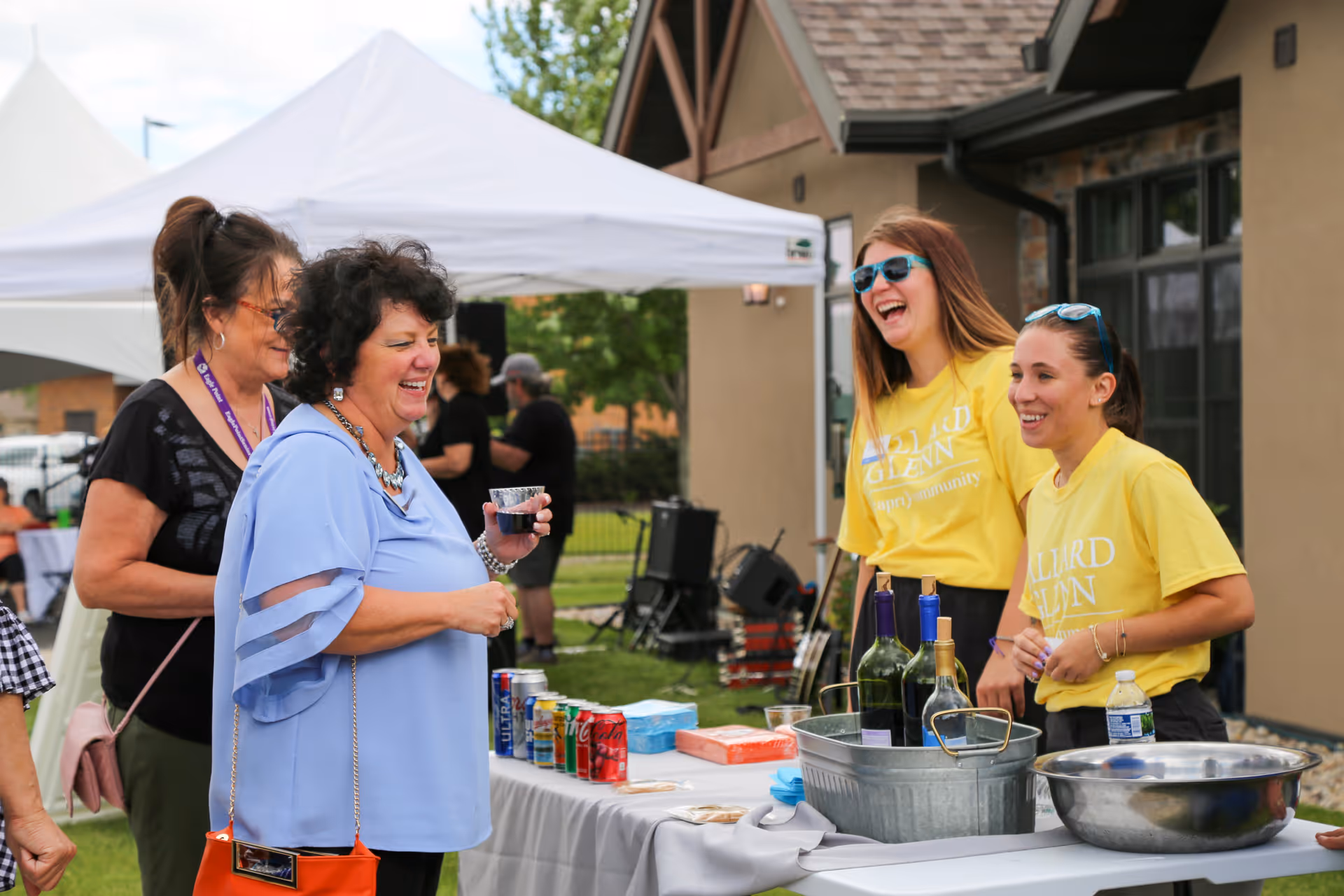 Two women wearing yellow Ballard Glenn t-shirts stand behind a table with drinks and snacks, smiling and interacting with two other women standing in front of the table at an outdoor event with a white canopy tent and a building in the background.