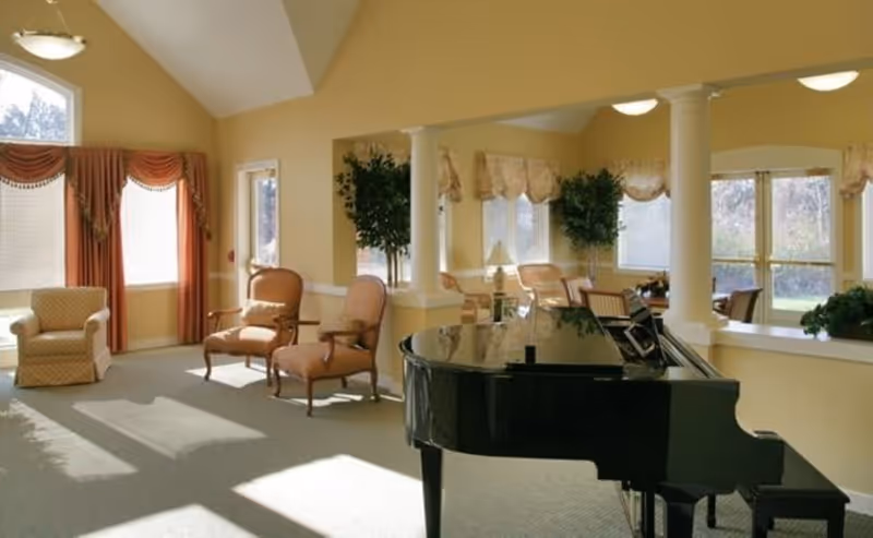 Bright senior living common room with a black grand piano, armchairs, columns, and large windows letting in sunlight.