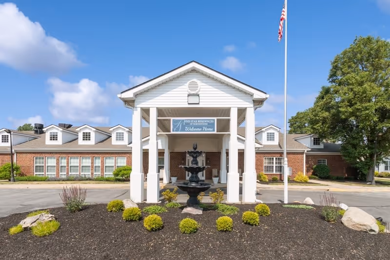 Front exterior view of Five Star Residences of Northwoods building with a covered entrance, a fountain in front, landscaped bushes, an American flag on a flagpole, and a clear blue sky with some clouds.