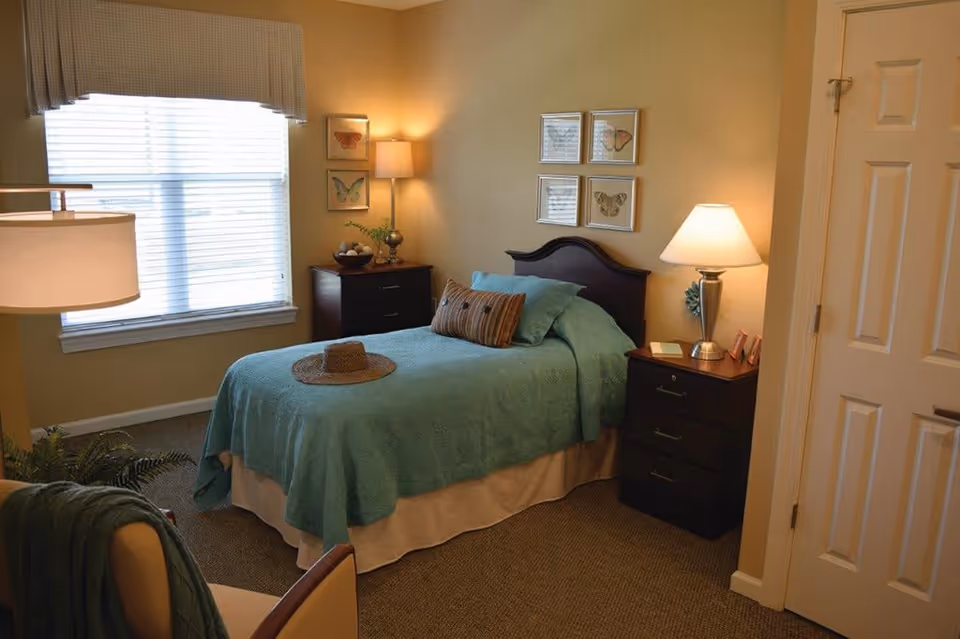 A cozy bedroom with a single bed covered in a teal quilt and pillows. There is a straw hat placed on the bed. The room has two dark wooden nightstands with lamps on each side of the bed. The walls are decorated with framed butterfly artwork. A window with blinds and a valance lets in natural light. A chair with a green throw is partially visible in the foreground.