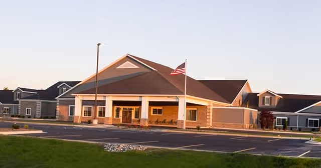 Exterior view of a single-story senior living facility building with a large covered entrance, an American flag on a flagpole, and a parking lot in front. The building has a brown roof and beige walls with white pillars supporting the entrance roof.