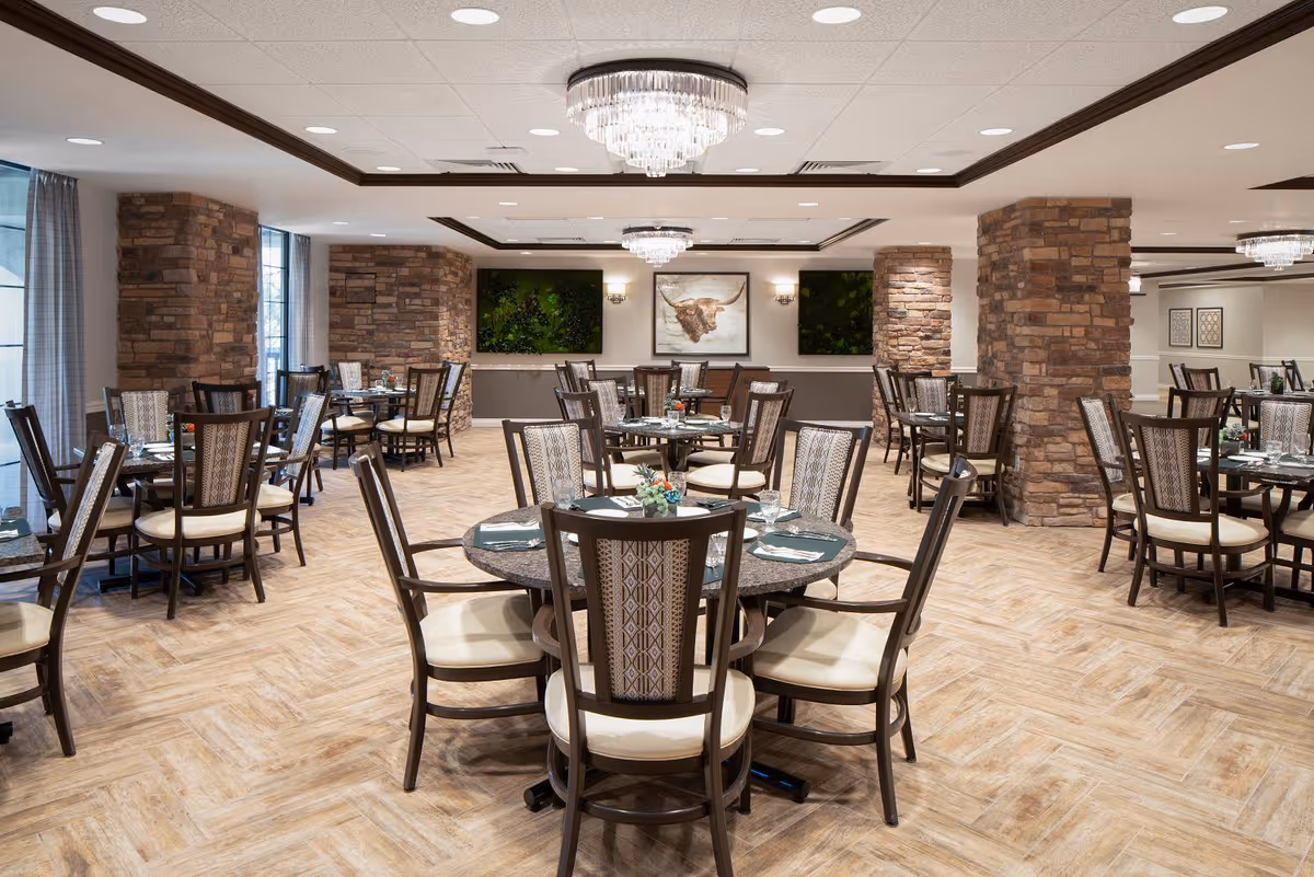 A spacious dining room with multiple round tables set with plates, glasses, and napkins. The room features wooden chairs with cushioned seats, stone pillars, large windows with curtains, and elegant chandeliers hanging from the ceiling. Artwork and greenery adorn the walls.