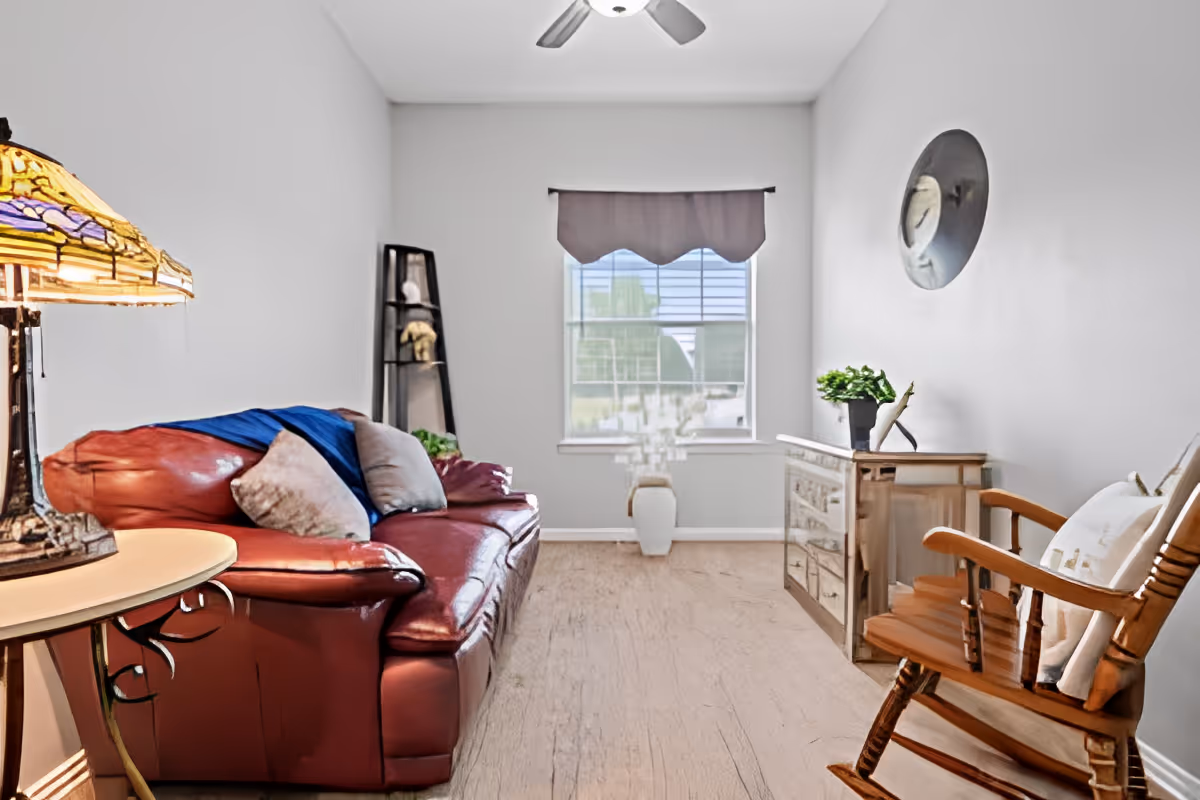 A cozy living room with a red leather sofa adorned with cushions and a blue throw blanket, a wooden rocking chair with a cushion, a decorative lamp on a small round table, a tall black shelf with decorative items, a wooden dresser with a plant and sculpture on top, a window with a valance, and a ceiling fan above.
