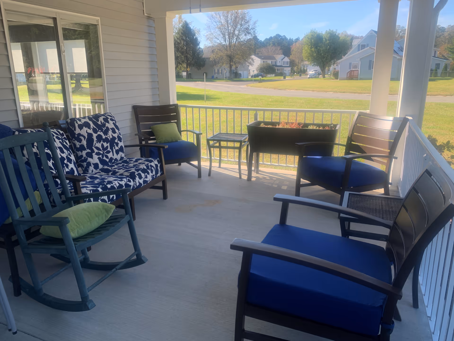 A covered outdoor porch area with several chairs including a blue rocking chair with a green pillow, a loveseat with blue and white floral cushions, and multiple armchairs with blue cushions. There is a small table and a planter box with plants. The porch overlooks a grassy area with houses and trees in the background under a clear sky.