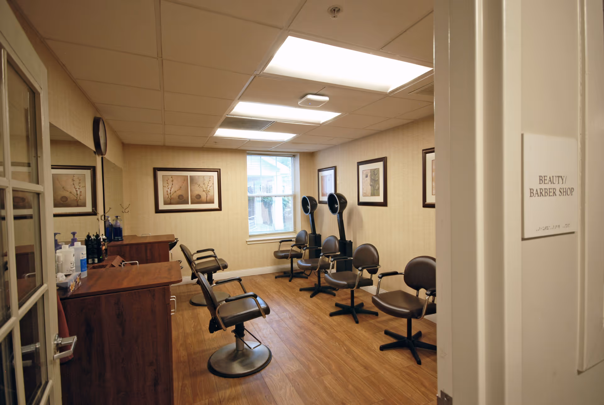 Interior view of a beauty and barber shop with several brown salon chairs, hair drying stations, wooden flooring, framed artwork on the walls, and a window letting in natural light.