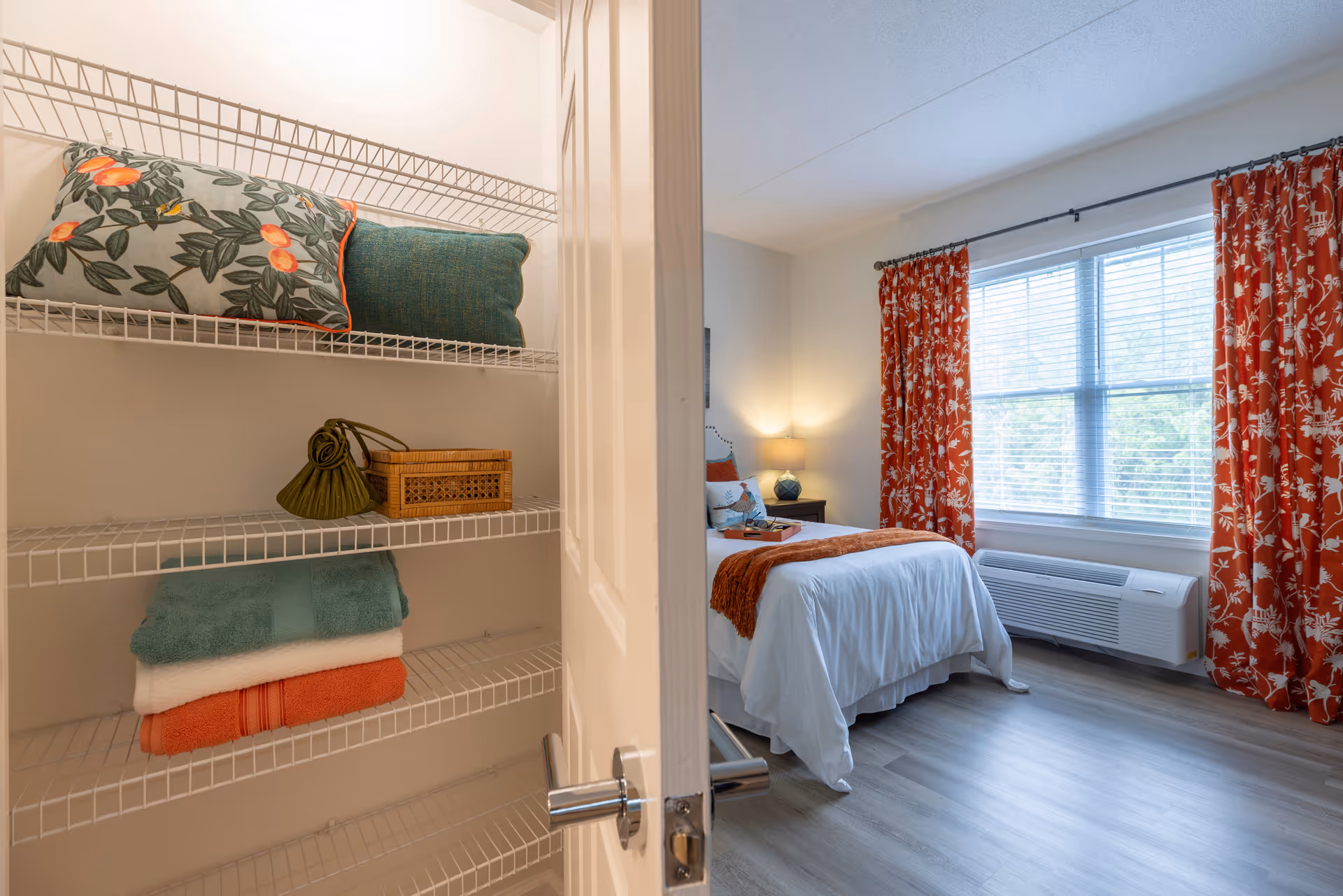 View through an open door into a bedroom with a neatly made bed featuring white bedding and an orange throw. The room has large windows with orange floral curtains and a bedside table with a lamp. In the foreground, there is a closet with wire shelves holding folded towels, pillows, and a small decorative basket.