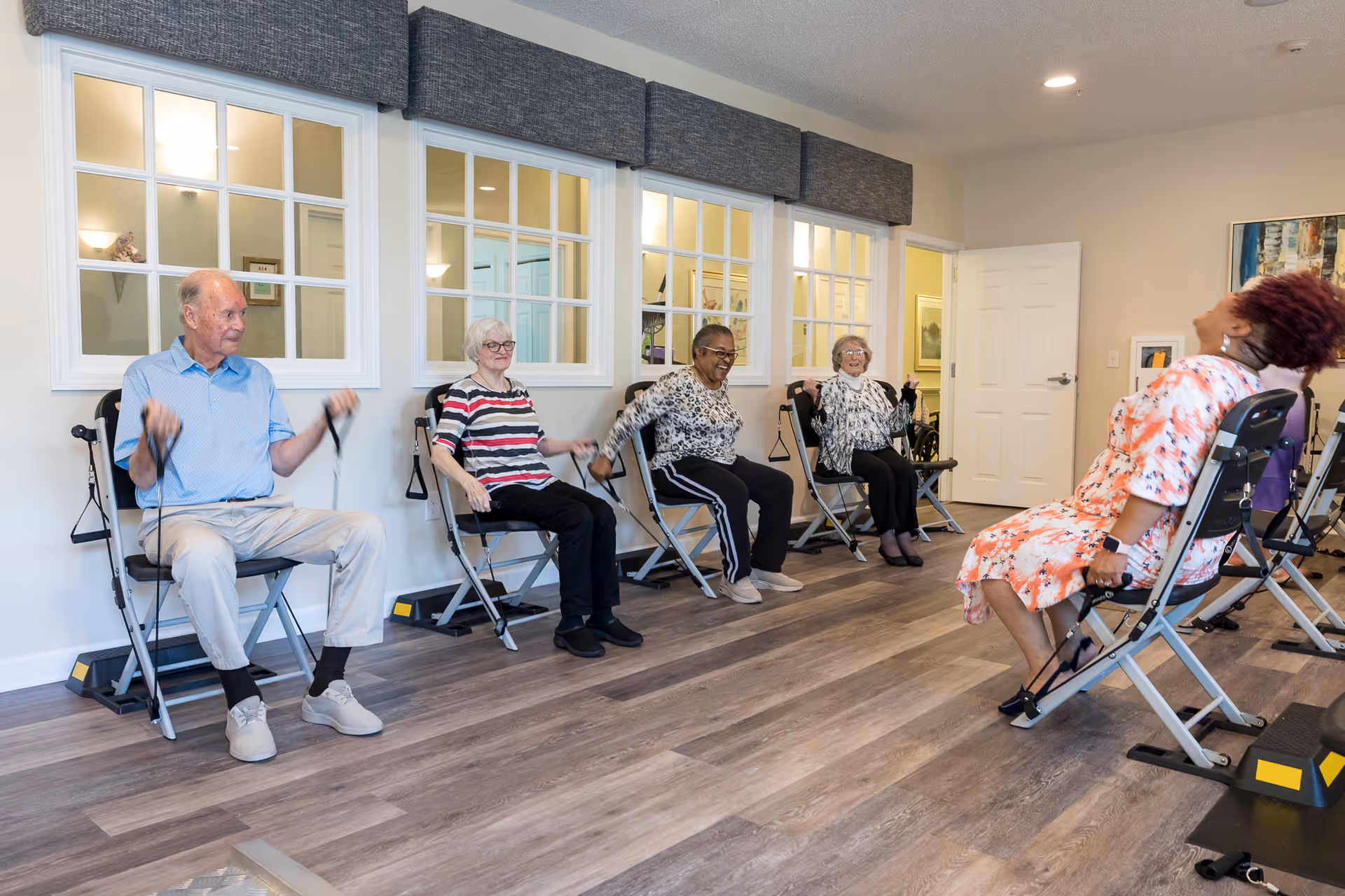 A group of five elderly people seated on exercise chairs in a bright room with wooden flooring, performing seated resistance band exercises. The room has large windows with white frames and a white door in the background.