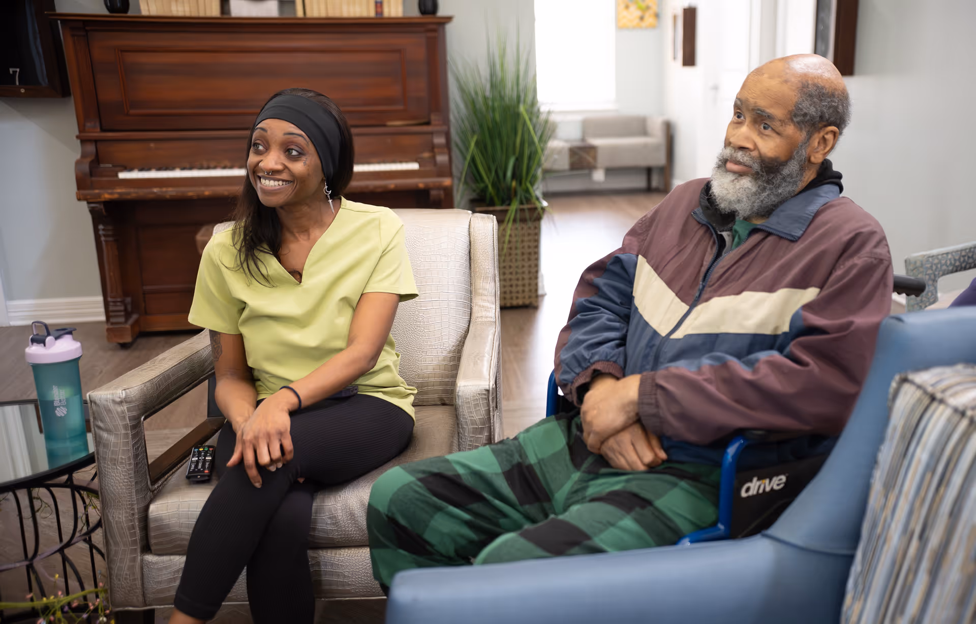 A smiling female caregiver in a light green scrub top sits on a chair next to an elderly man with a gray beard wearing a jacket and plaid pants, who is seated in a wheelchair. They are in a cozy room with a piano, a plant, and comfortable seating in the background.