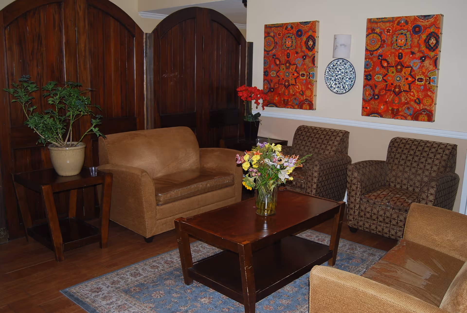 A cozy living room with a brown loveseat, two patterned armchairs, and a brown armchair arranged around a wooden coffee table with a vase of colorful flowers. There is a side table with a potted plant and a dark wooden folding screen in the background. Two vibrant red and orange patterned artworks and a decorative plate are mounted on the beige wall.