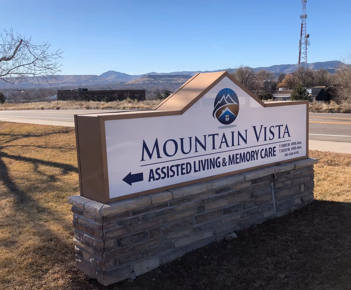 Outdoor sign for Mountain Vista Assisted Living & Memory Care with an arrow pointing left, set on a stone base with a road, trees, and mountains in the background under a clear blue sky.